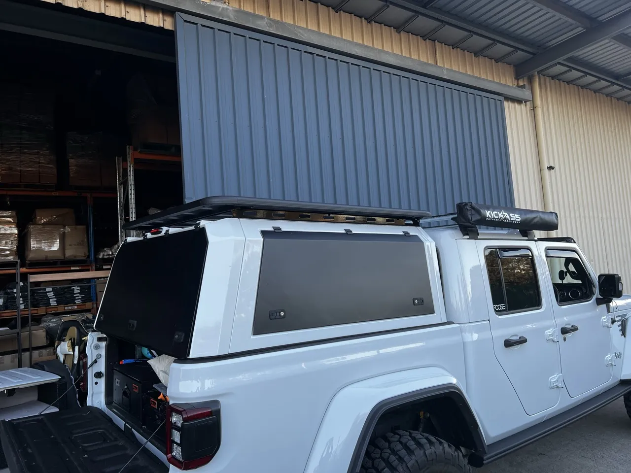 A Jeep Gladiator with a canopy and roof rack outside an open warehouse door, stock on the racks behind